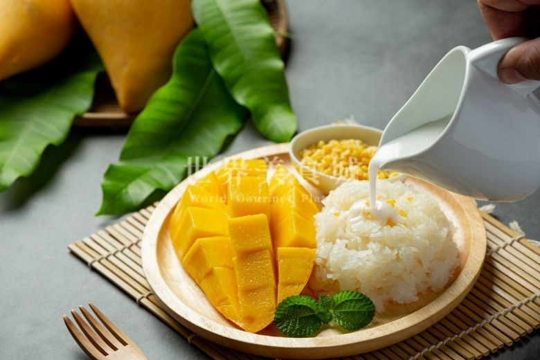 coconut milk pours onto mango sticky rice put on a wooden plate with mangoes in the background